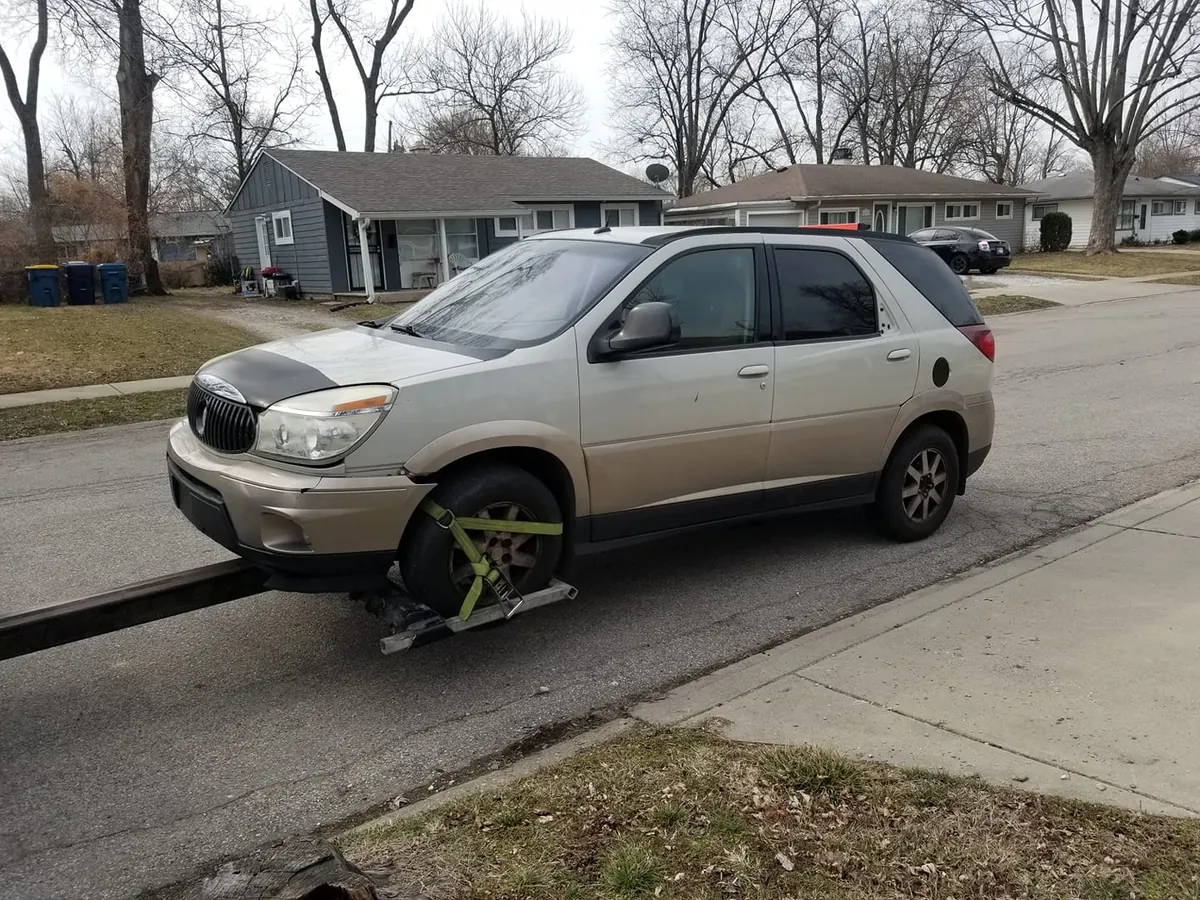 Two-tone Buick Rendezvous with front bumper damage being towed on an Indianapolis street, an SUV priced in the low junk car tier