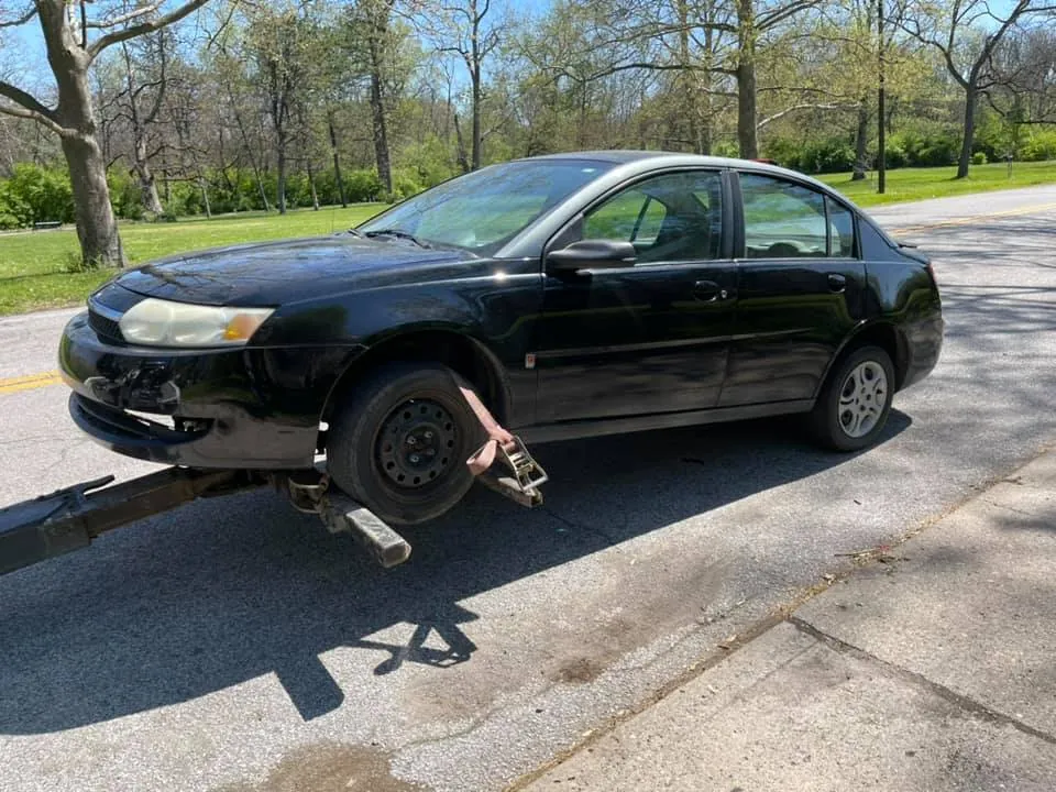 Sell a totaled car in Indiana: black sedan with a detached front bumper lifted by a tow truck for a cash pickup